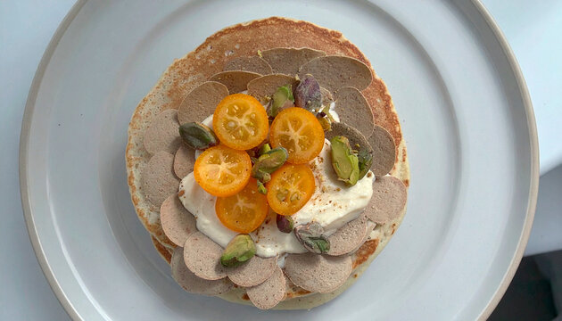 Stack of fluffy pancakes with whipped cream, kumquats, and pistachios in sunlight