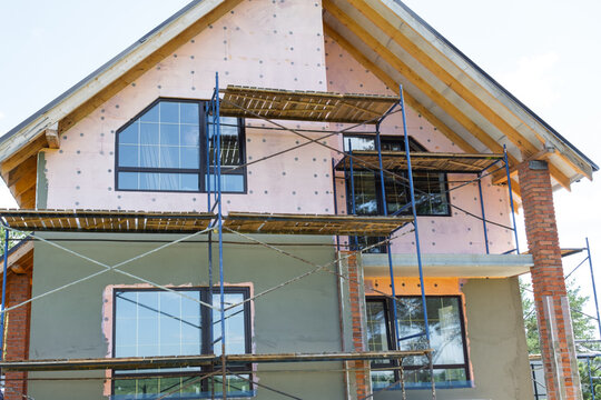 House under facade plastering, exterior scaffolding for render application, insulation layer, surface leveling, construction wall finish, decorative coating.