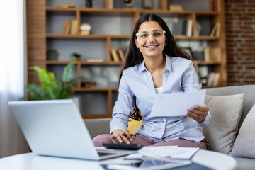 Fototapeta premium Young indian woman on a cozy living room sofa managing taxes and personal finances with laptop and paperwork, smiling confidently while budgeting and planning investments