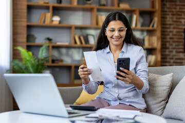 Fototapeta premium Young indian woman checking receipts on smartphone, doing calculations for taxes or budget planning, using modern digital banking and accounting tools at home