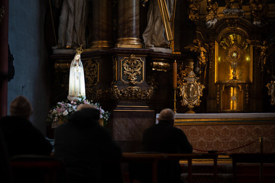 Devout people praying in a dim baroque church before the Blessed Sacrament and a statue of the Virgin Mary in Trzebnica, Poland.