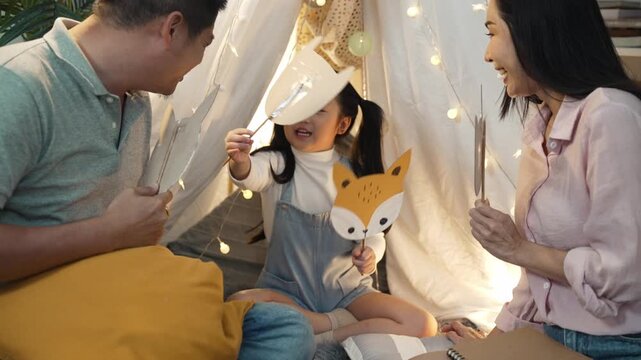 Happy Asian family with father, mother and daughter playing with animal paper masks in a cozy teepee tent at home. They are smiling and bonding together during storytelling time in a warm bedroom