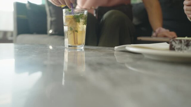 A glass of iced drink with mint leaves and a straw sits on a marble table in soft lighting, with blurred people in the background, creating a cozy, refreshing scene