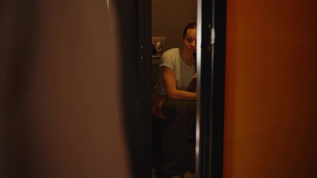 A woman sits on the toilet in a hotel bathroom, talking on the intercom, with a modern black sink visible in the foreground, capturing a candid and unusual moment