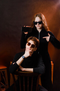 Cool teenagers posing in a dark studio, girl showing a black mobile phone screen and pointing at a sitting guy.
