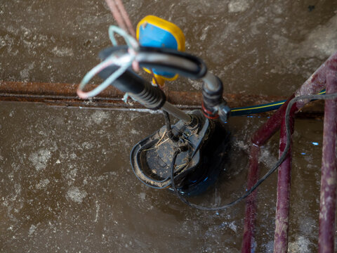 Black electric submersible water pump with yellow float switch operates in muddy brown wastewater to drain flooded residential basement or construction site pit.