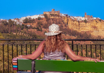 View from behind of a woman with a straw hat sitting on a bench at the Mirador de Abades, contemplating the white town of Arcos de la Frontera with the castle of the Dukes of Arcos in Cadiz, Spain.