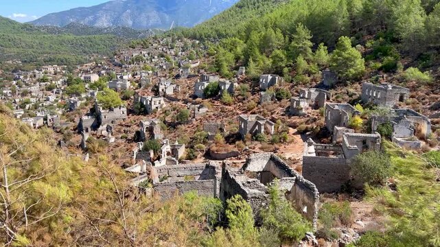 Abandoned stone houses scattered across hillside of Kayakoy ghost village near Fethiye, Turkey, surrounded by pine forest and mountains under clear summer sky, historic deserted settlement landscape