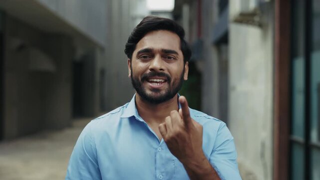 Portrait of happy Indian man in blue shirt using sign language on old city street. Young bearded man smiling and saying Hello, how are you. Deaf communication concept.