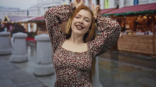 Redhead woman showing bare decolletage and hands on head making bunny ears gesture, tongue out, in a busy street market; playful spontaneity.