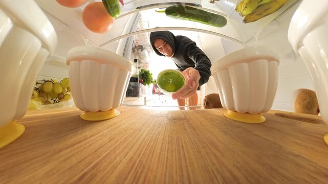 View from inside a refrigerator as a man opens the door and contemplates between green and red jelly cups. The wide-angle shot captures various foods on a wooden shelf in the foreground