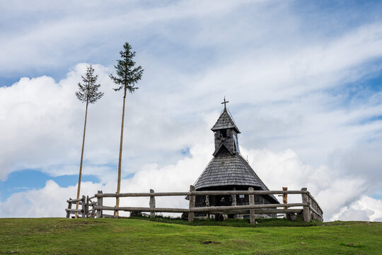 A wide mountain landscape showing a brown cow grazing in the foreground while hikers walk up a path toward a wooden chapel on a green hill in Slovenia.