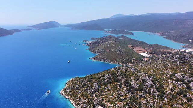 Aerial panoramic view of Kekova island coastline with excursion boats sailing turquoise waters surrounded by forested hills and distant mountains, Antalya, Turkey