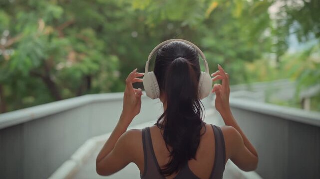 Rear view of cheerful asian woman wearing headphones and preparing to run in public park during morning &ndash; healthy lifestyle, wellness motivation, and outdoor fitness routine with peaceful nature.