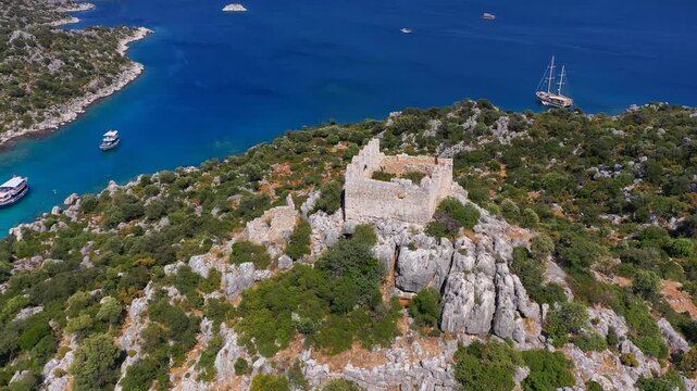 Aerial view of Byzantine castle ruins on rocky hilltop at Tersane Island with excursion boats anchored in turquoise cove below, Kekova, Antalya, Turkey