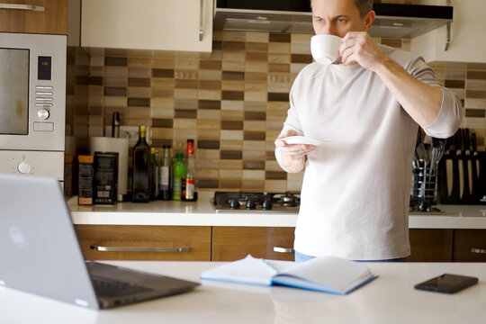 Man Drinking Coffee While Working Remotely in Modern Kitchen. Man standing in a contemporary kitchen, drinking from a white cup while looking at laptop. Home office scene with notebook and smartphone