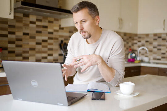 Man Having Video Call and Gesturing at Laptop in Modern Kitchen. A man sitting at a kitchen, gesturing with his hands during an online meeting. Home office setting with a laptop, notebook, and coffee