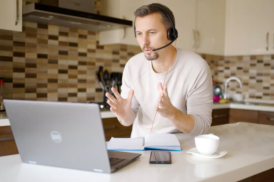 Man in Headset Having Online Meeting in Modern Kitchen. Focused man wearing a headset with microphone while talking during a video conference on his laptop. Home office and remote work