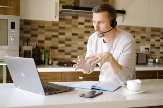 Man in Headset Having Online Meeting in Modern Kitchen. Focused man wearing a headset with microphone while talking during a video conference on his laptop. Home office and remote work