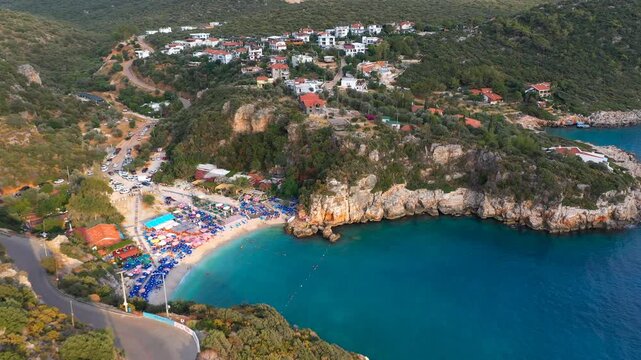 Aerial view of Buyuk Cakil Beach with colorful umbrellas in sheltered cove surrounded by rocky cliffs and hillside village, Kas, Antalya, Turkey