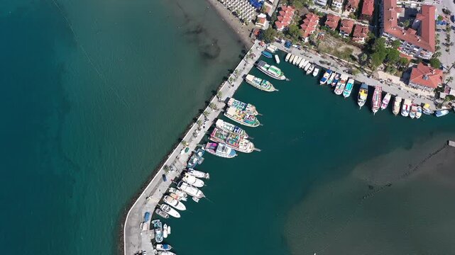 Aerial top-down view of Akyaka resort town with colorful tour boats moored at pier, sandy beach with umbrellas and red-roofed buildings, Mugla, Turkey