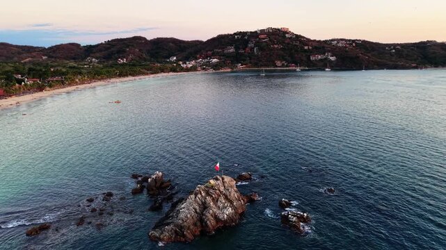 Aerial drone orbit of Mexican flag waving on rocky outcropping in Pacific Ocean with Playa La Ropa beach bay and hillside resort town of Zihuatanejo, Mexico at golden hour