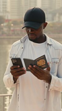 Man in baseball cap types on phone screen while holding British passport. African American guy sends document details for verification via smartphone standing on city embankment