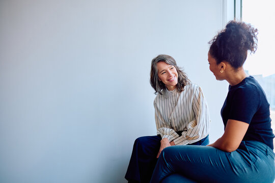 Two diverse businesswomen smiling during an informal meeting, engaging in a pleasant conversation about work in a bright workplace