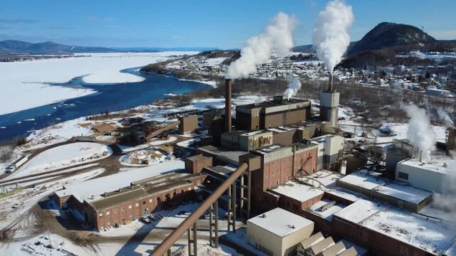 Cinematic aerial shot of an industrial plant by a frozen river in New Brunswick. Steam rises from chimneys against a backdrop of snowy mountains and a quiet town. Cold energy production scene.
