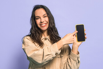 Fototapeta premium A woman stands in a studio with a purple background. She smiles while pointing at her smartphone. The device has a yellow case. Her hair is long and wavy.