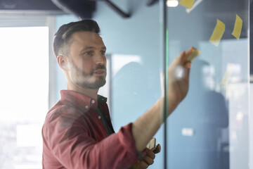 A man is writing on a glass board in an office. He is focused on arranging sticky notes while...