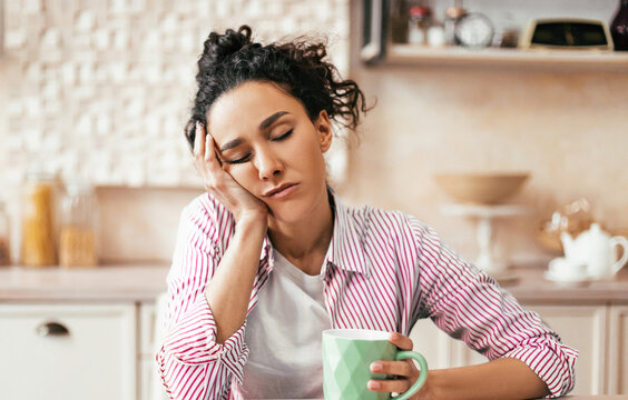 A woman sits at a kitchen table with a green mug in one hand and a tired expression on her face. Cookies are placed on the table nearby. The kitchen is simple and modern.