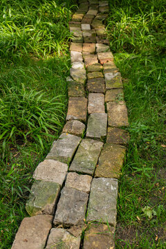 Narrow straight path made of irregular old weathered stone and brick pavers running through lush green grass, top down perspective.

