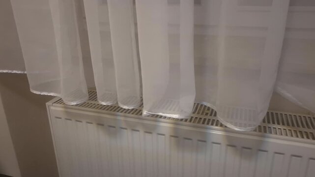Close-up of curtains gently hanging above radiator near window in soft indoor lighting. Static camera captures minimal interior scene with calm atmosphere and natural light.