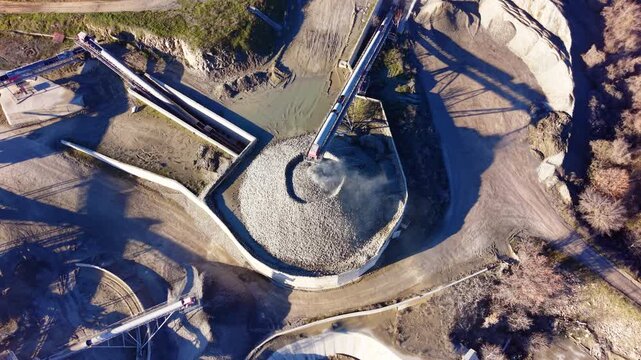 Large circular pile of crushed stone being fed by a long conveyor belt in a processing plant. Aerial top view of circular stone stockpile and conveyor system.