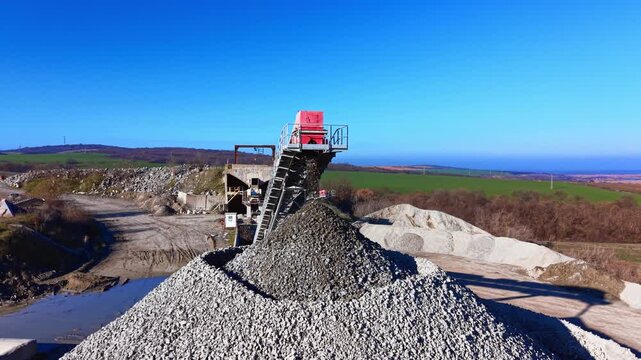 Crushed stone falling from the end of a high conveyor onto a massive pile against a clear blue sky. Aerial view of gravel falling from conveyor belt.