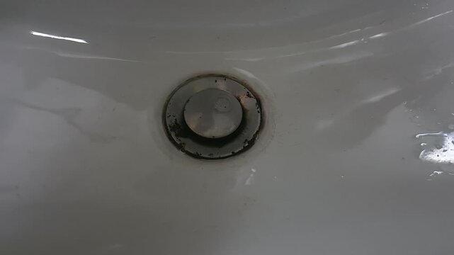 Close-up of water flowing from faucet into sink drain. Static camera captures stream hitting basin and creating splashes in clean bathroom environment.