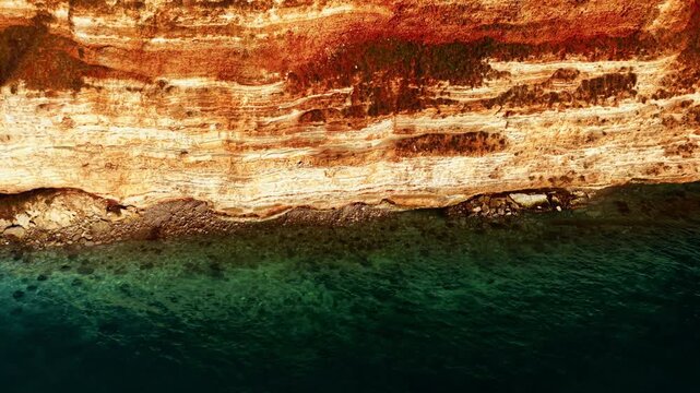 Aerial close up of sedimentary rock layers on a cliff face meeting green sea water. Layered orange limestone sea cliffs.