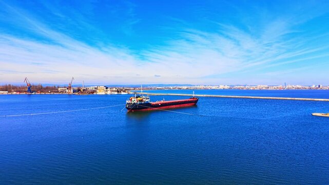Large bulk carrier vessel stationary in open water with a city skyline and port in the distance. Cargo ship anchored in blue sea near breakwater wall.