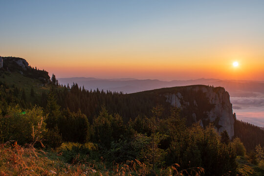 Sunset over Ceahlau Massif near Cabana Dochia, Romania, with forested ridges and rocky cliffs above a sea of clouds