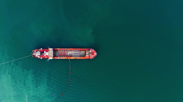 Directly overhead shot of a red industrial vessel in dark green water connected to a floating pipeline with buoys. Top view of red dredging ship pumping sand through pipe.