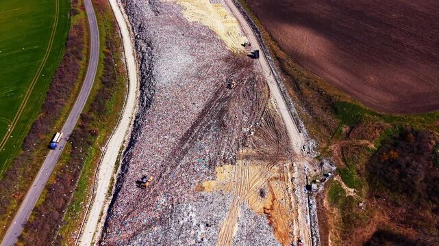 Long landfill area adjacent to a paved road and green cultivated farmland. Large scale waste disposal site near agricultural fields.