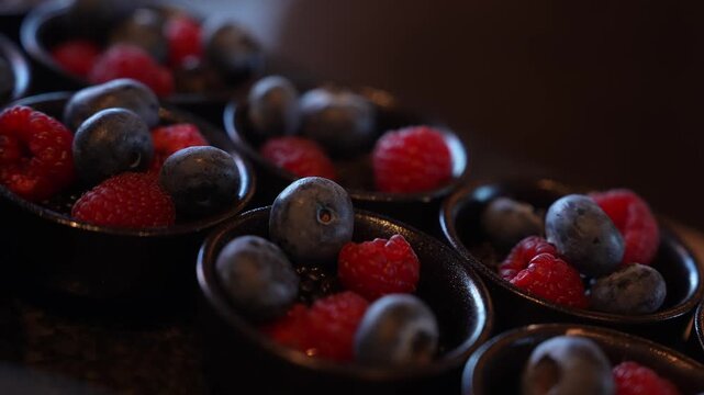 Rows of small black ceramic tasting cups hold raspberries and blueberries on a dark stone surface. The camera glides slowly with shallow depth of field and soft bokeh.