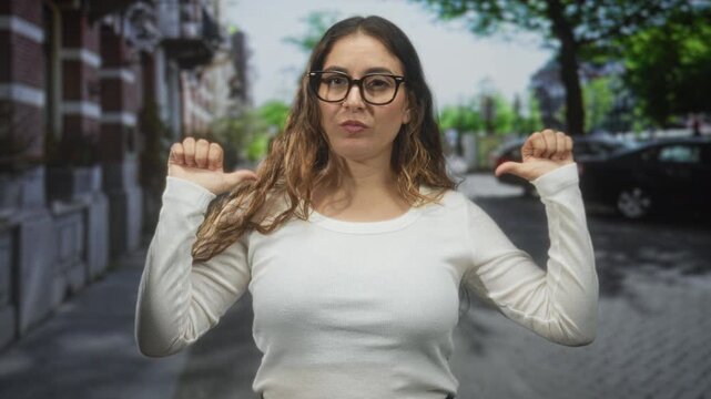 Woman thumbs pointing to self on urban street with parked cars and brick building, glasses and white shirt visible; confidence.