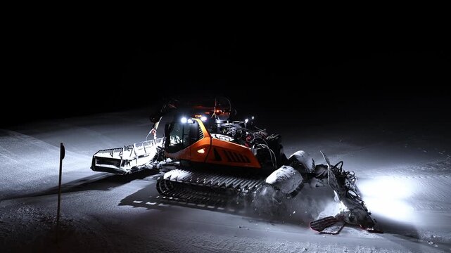 Orange tracked PistenBully style groomer smooths fresh snow at an Alpine ski resort at night, LED lights shine, corduroy patterns form, chalet glows by the slope edge