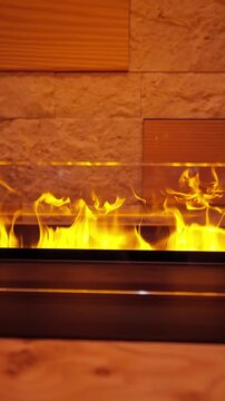 Vertical view of a modern linear fireplace as yellow flames rise behind a glass guard, framed by rough stone and wood panels in an Alpine lodge, evening winter scene.