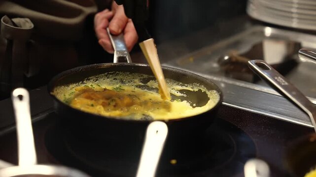 A chef in black gloves folds herb scrambled eggs in a black skillet as steam rises, with plates and stainless work surfaces in a pro kitchen, warm and shallow DOF.