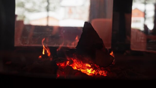 Low angle close up shows a charred log, molten coals, and gentle flames in a wood stove. Snow dusted rooftops and pine trees sit outside, evoking an Alps winter.