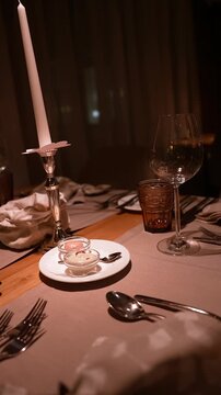 A candlelit place setting sits on a wooden table with a white taper, a wine glass, an amber tumbler, ramekins, cutlery, and linens as the camera pans slowly.