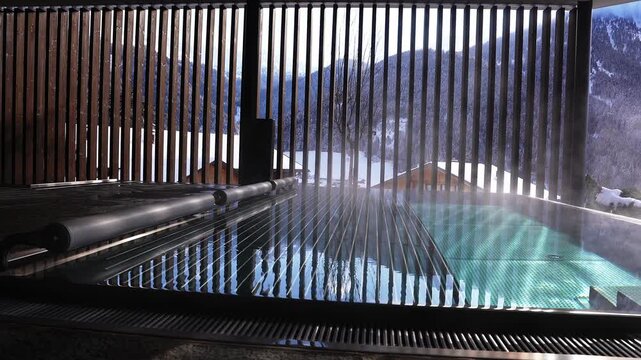 Steaming heated plunge pool with retractable cover on chalet terrace, framed by vertical wooden slats, overlooks snowy rooftops and Alpine slopes in winter.